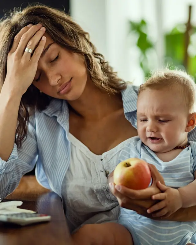 Mãe cansada, com a mão na testa, segurando um bebê que chora diante de uma maçã