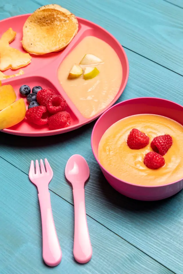 Pratinho rosa com papinha, panqueca e frutas vermelhas, ao lado de um potinho de purê
